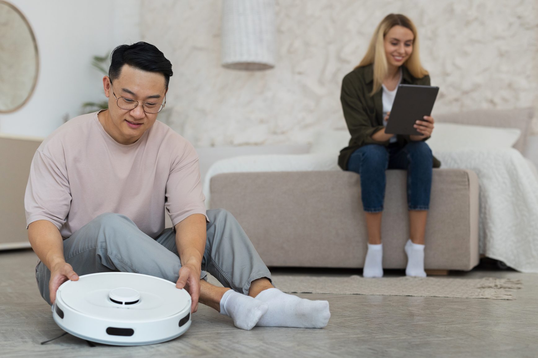 Man setting up a robot vacuum on floor while woman uses tablet on sofa in modern living room