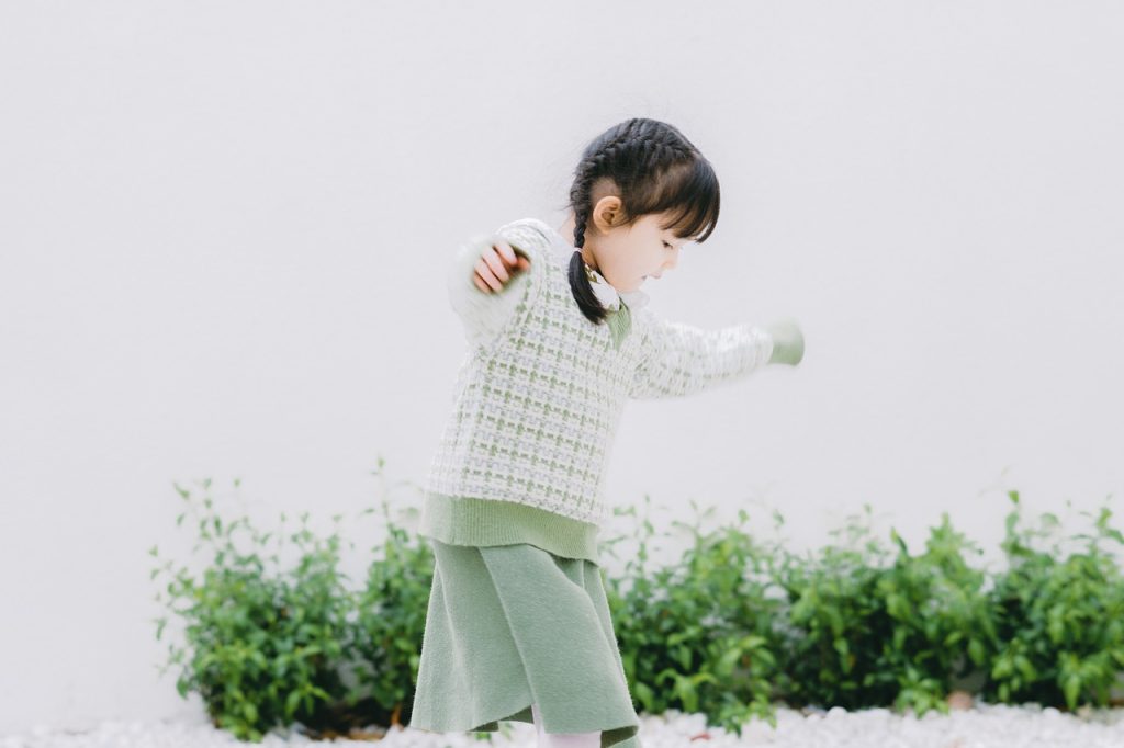 A young girl wearing a green sweater and skirt, smiling joyfully in a bright outdoor setting
