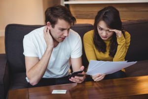 A man and woman sit on a couch looking worried while examining papers and a smartphone, possibly reviewing bills or financial documents.