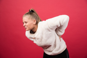 A woman stands against a red background, holding her back in discomfort